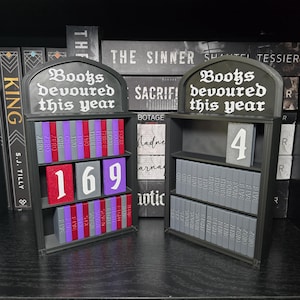May include: Two black 3D-printed bookshelf trackers with the text Books devoured this year at the top. The left unit displays the number 169 using red, grey, and purple book blocks. The right unit displays the number 4 using grey book blocks.