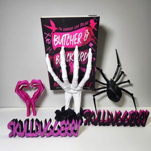 May include: A book titled "Butcher & Blackbird" with a black cover and pink accents, propped up by a white skeleton hand. A pink heart-shaped sculpture and a black spider are also present, along with a pink and black word art piece that reads "SkullDuggery".
