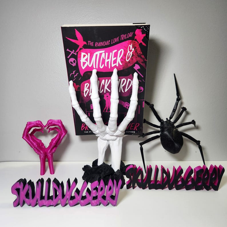 May include: A book titled "Butcher & Blackbird" with a black cover and pink accents, propped up by a white skeleton hand. A pink heart-shaped sculpture and a black spider are also present, along with a pink and black word art piece that reads "SkullDuggery".