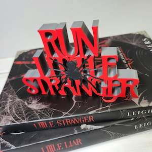 Puede incluir: Un sujetalibros impreso en 3D con las palabras "RUN LITTLE STRANGER" en rojo y gris, con una ara&ntilde;a negra en el centro. El sujetalibros descansa sobre dos libros con el t&iacute;tulo "LITTLE STRANGER".