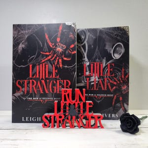 Puede incluir: Dos libros titulados "Little Stranger" con un gr&aacute;fico de ara&ntilde;a roja y un letrero de madera rojo que dice "Run Little Stranger". Una rosa negra est&aacute; a la derecha. Los libros son parte de "The Web of Silence Duet".