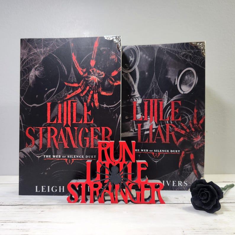 Puede incluir: Dos libros titulados "Little Stranger" con un gr&aacute;fico de ara&ntilde;a roja y un letrero de madera rojo que dice "Run Little Stranger". Una rosa negra est&aacute; a la derecha. Los libros son parte de "The Web of Silence Duet".