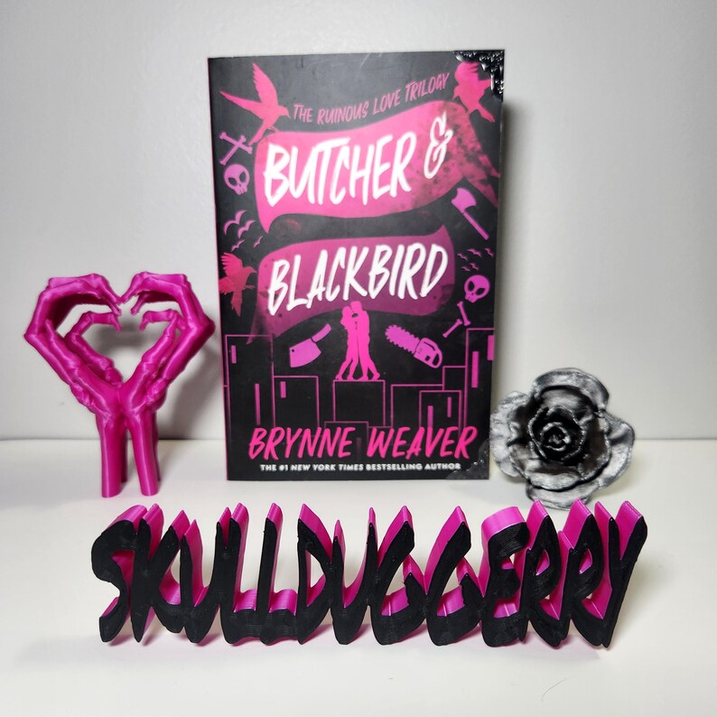 May include: A book titled "Butcher & Blackbird" by Brynne Weaver, with a black and pink cover, is displayed with a pink hand-shaped sculpture, a black and white rose, and a "SKULLDUGGERY" word art piece.