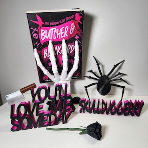 May include: A book titled "Butcher & Blackbird" with a skeleton hand, a black spider, and the phrases "You'll Love Me Someday" and "Skullduggery" in black and pink. A small cleaver and a black rose are also present.