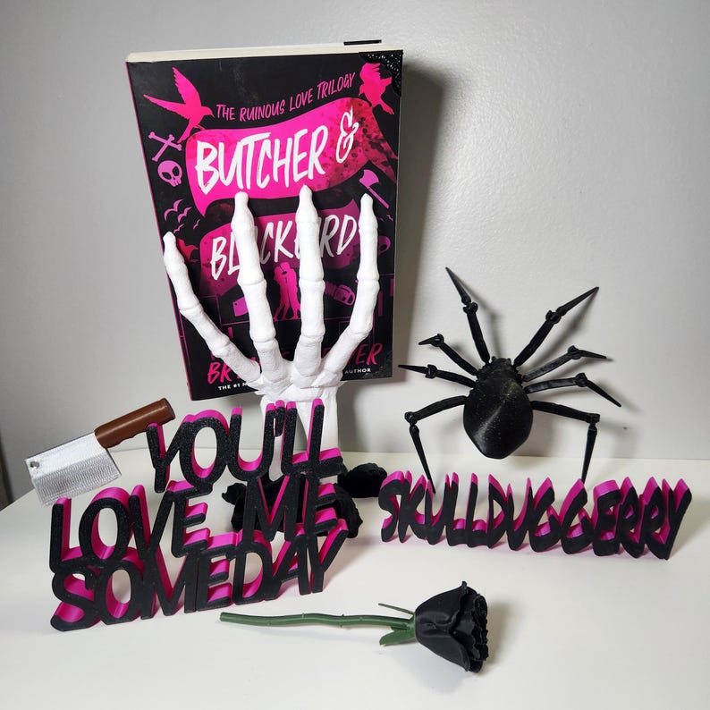 May include: A book titled "Butcher & Blackbird" with a skeleton hand, a black spider, and the phrases "You'll Love Me Someday" and "Skullduggery" in black and pink. A small cleaver and a black rose are also present.