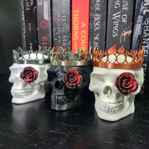May include: Three skull-shaped decorative planters. One is white, one is black, and one is off-white. Each skull has a red rose over one eye and a crown. The crowns are silver, gold, and copper-colored. Books are in the background.