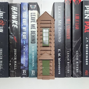 May include: A brown, vertical, log-cabin-style book nook insert featuring a green door at the base and a green-framed window above. The structure is positioned between several hardcover books with black, white, and blue spines on a shelf.