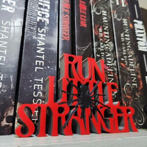Puede incluir: Un sujetalibros rojo y gris impreso en 3D con las palabras "RUN LITTLE STRANGER" y una ara&ntilde;a negra. El sujetalibros est&aacute; colocado frente a una fila de libros con cubiertas oscuras.