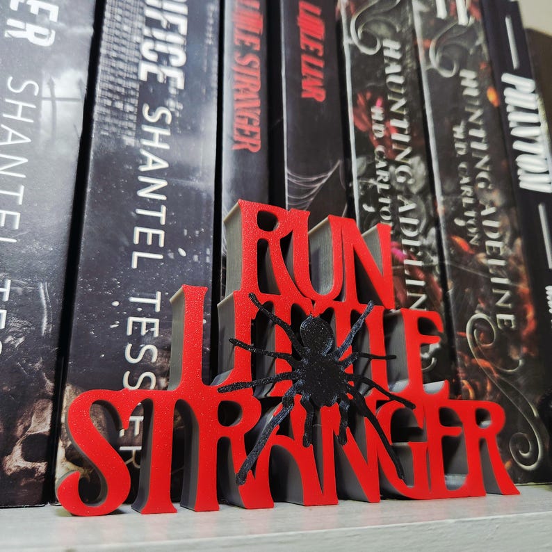 Puede incluir: Un sujetalibros rojo y gris impreso en 3D con las palabras "RUN LITTLE STRANGER" y una ara&ntilde;a negra. El sujetalibros est&aacute; colocado frente a una fila de libros con cubiertas oscuras.