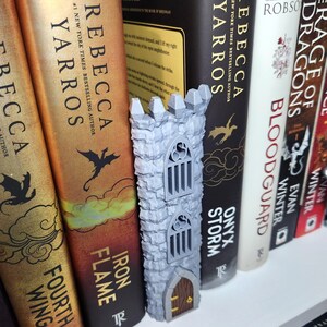May include: A gray 3D-printed tower figurine with a stone-textured exterior, featuring two arched windows with vertical bars and a small brown arched door at the base. The tower is positioned vertically between several hardcover books on a shelf.