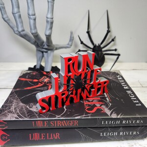 Puede incluir: Una exhibici&oacute;n tem&aacute;tica de Halloween con un letrero rojo y gris impreso en 3D que dice "RUN THE STRANGER" con un acento de ara&ntilde;a negra. El letrero est&aacute; encima de dos libros con dise&ntilde;os de telara&ntilde;as. Una mano de esqueleto gris y una ara&ntilde;a negra est&aacute;n en el fondo.