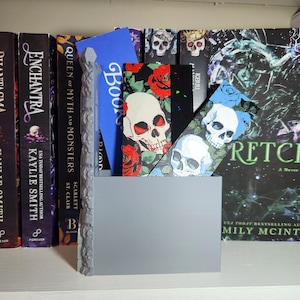 May include: A gray rectangular holder with a textured stone-like pillar on the left side, containing several bookmarks. The bookmarks feature designs including a blue one with the word Book, a black one with red roses and a skull, and a blue floral skull.