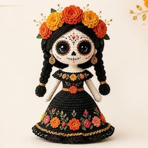 Puede incluir: Una muñeca de ganchillo con cara blanca, marcas faciales negras tipo calavera, ojos negros grandes y dos largas trenzas negras. La muñeca viste un vestido negro con detalles florales bordados y una corona de flores naranjas y amarillas.