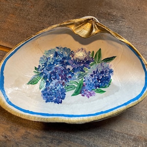 May include: A white seashell painted with a blue and purple floral design. The shell has a gold rim and a blue band around the edge.