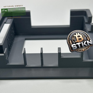 May include: A gray plastic case with a rectangular opening and four vertical dividers. The case has a textured surface and is labeled "Veteran Owned" with a US flag graphic. A gold coin with a Bitcoin symbol and the text "STKN" is visible in the background.