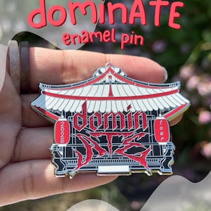 May include: A red and black enamel pin with the word "domiNATE" in red script above the pin. The pin features a pagoda design with the words "dominATE" and "2023 tour" in red. The pin is held by a person.