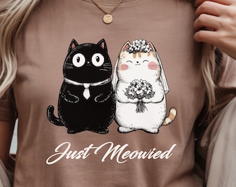 May include: An espresso-colored t-shirt featuring a graphic of two cats dressed for a wedding. The black cat wears a white tie, and the white cat wears a floral veil and holds a bouquet. Below the cats, the text reads Just Meowied in white script.