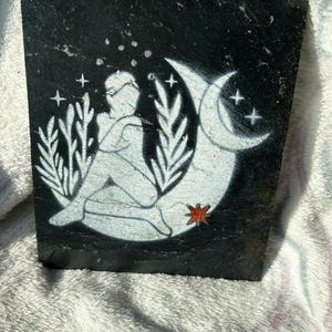 Może przedstawiać: Czarny podkładka ze łupka z białym wzorem namalowanym osoby siedzącej na półksiężycu z gwiazdami i czerwoną gwiazdą.
