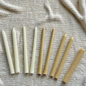 May include: Assortment of eight candles in various sizes and styles. The candles are made of beeswax and have a natural, off-white color. Some candles have a ribbed texture, while others are smooth. The candles are arranged in a row on a beige fabric background.