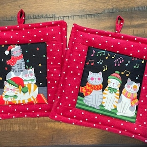 May include: Two square fabric pot holders with red and white polka dot borders and fabric hanging loops. The center panels feature black backgrounds with colorful illustrations of cats wearing winter scarves and hats, with musical notes.