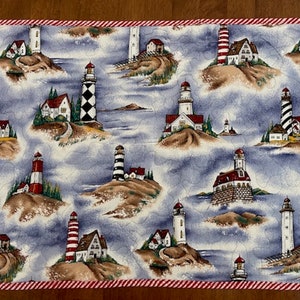 May include: A blue and white quilted table runner with a lighthouse and seascape design. The runner has a red and white striped border.