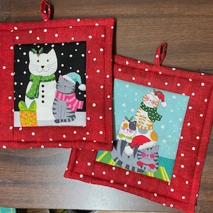 May include: Two red quilted pot holders with white polka dots. One pot holder features a snowman and a cat wearing a pink scarf and a blue hat. The other pot holder features three cats stacked on top of each other, wearing Santa hats and scarves.