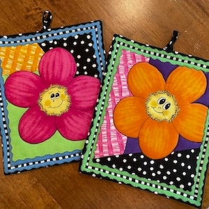 May include: Two colorful pot holders with a flower design. Each pot holder has a smiling face in the center of the flower. One pot holder has a pink flower with a green, orange, and black background. The other pot holder has an orange flower with a purple, pink, and black background.