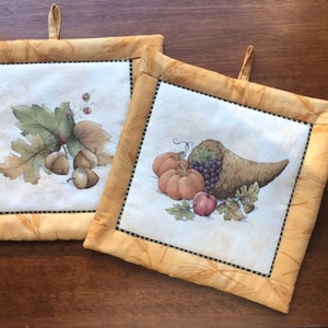 May include: Two square pot holders with a fall theme. One features acorns and leaves, the other a cornucopia with pumpkins, grapes, and an apple. Both have a yellow border with a wheat pattern and a black dotted trim.