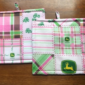 May include: Two square pot holders with a patchwork design. The pot holders feature pink, green, and brown plaid patterns, with green tractor graphics and the John Deere logo. Each pot holder has a small fabric loop for hanging.