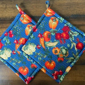 May include: Two square fabric pot holders featuring a blue background with a woven texture pattern. The surface is decorated with a colorful print of various vegetables including red tomatoes, orange carrots, yellow corn, and white cauliflower.