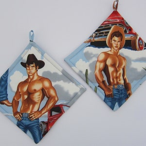 May include: Two diamond-shaped pot holders with a cowboy theme. Each pot holder features a muscular man in a cowboy hat and jeans, set against a backdrop of a red truck and a blue sky with clouds. The pot holders have a light blue border and a loop for hanging.