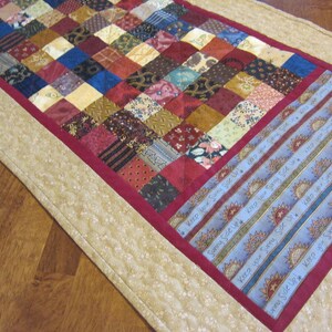 May include: A patchwork table runner with a red border and a beige outer border. The runner is decorated with blue and white fabric strips with the text "Keep your Sunny Side Up" repeated on each strip.
