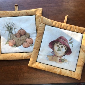 May include: Two square pot holders with a fall theme. One features pumpkins on a hay bale, the other a scarecrow. Both have a cream-colored center with a black border and a gold wheat-patterned border.
