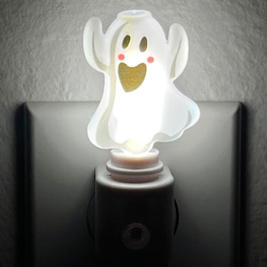 May include: A white ghost-shaped night light with a smiling face, rosy cheeks, and a golden heart-shaped mouth. The night light is plugged into a wall outlet, providing a soft, warm glow.