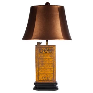 May include: A table lamp featuring a metallic bronze-colored lampshade and a base constructed from a rectangular, weathered yellow O-Cedar Polish metal tin. The lamp sits on a tiered black square base and includes a small finial on top.
