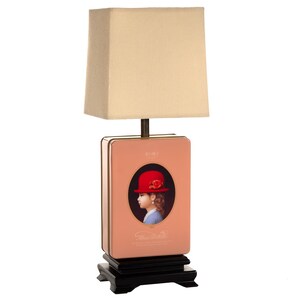 May include: A decorative table lamp with a rectangular peach-colored base featuring a portrait of a girl in a red hat. The lamp has a beige square lampshade and sits on a black wooden base.