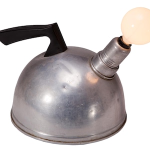 May include: A repurposed metal kettle lamp with a black handle and a light bulb. The kettle is silver-colored and has a rounded shape. The light bulb is round and emits a warm glow. The lamp is a unique home decor item.
