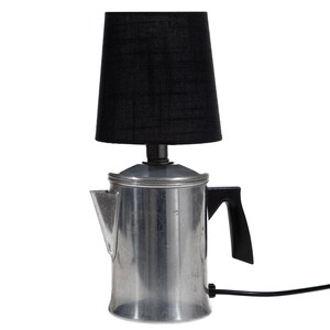 May include: A table lamp constructed from a silver-colored metal coffee percolator base with a black handle and a black power cord. The lamp features a cylindrical black fabric lampshade positioned directly on top of the metal percolator lid.