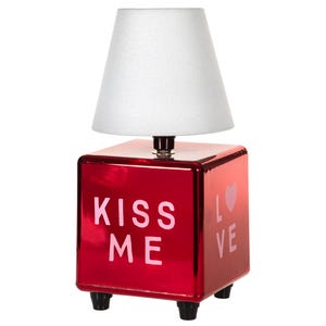 May include: A table lamp with a white conical shade and a reflective red cube base. The front of the base features the pink text KISS ME, while the side displays the pink text LOVE with a small pink heart icon. The base stands on four small black feet.