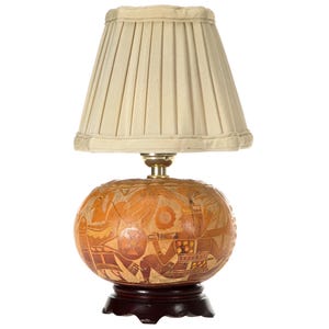 May include: A table lamp featuring a round, light brown gourd base with intricate, hand-carved figurative designs in darker brown tones. The lamp sits on a dark brown, scalloped wooden base and is topped with a cream-colored, pleated fabric shade.