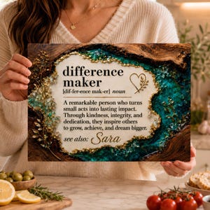 以下が含まれることがあります： 中央に白いパネルがあり、「difference maker [dif-fer-ence mak-er] noun」というテキストと定義が記された長方形の木製プレート。周囲は濃い木目の縁取りで、ターコイズブルーとゴールドの樹脂のアクセントとハートのアイコンがあしらわれています。
