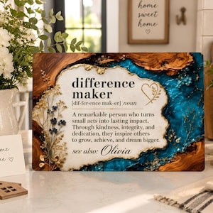 May include: A rectangular wooden plaque with a central white resin inlay featuring the text: difference maker [dif-fer-ence mak-er] noun. A remarkable person who turns small acts into lasting impact. see also: Olivia. The border has blue resin and gold leaf.