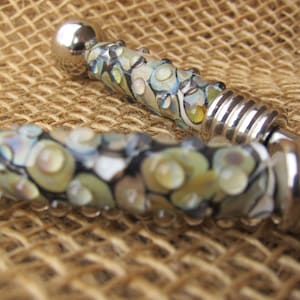 Puede incluir: Dos primeros planos de cuentas de vidrio decorativas con detalles plateados. Las cuentas son de vidrio y tienen una variedad de colores, incluyendo blanco, negro y azul. Las cuentas están enhebradas en un alambre plateado.