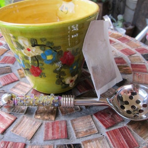 May include: A green ceramic teacup with floral designs, a tea bag, and a silver tea infuser with a glass handle on a tiled surface.