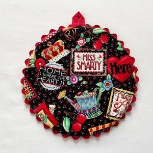 May include: Round, black fabric potholder with a red scalloped edge and a red hanging loop. The potholder is decorated with various images, including cherries, flowers, and the text "MISS SMARTY" and "HOME IS WHERE THE HEART IS".