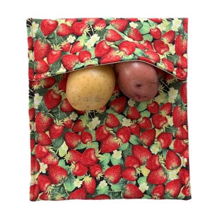 Può includere: Una borsa riutilizzabile per la spesa in tessuto, con un motivo a fragole rosse e foglie verdi, contenente una patata gialla e una rossa. La borsa ha una chiusura a patta. Lo sfondo è bianco.