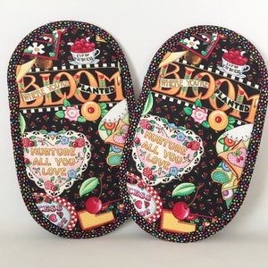 Pode incluir: Um par de pegadores de panela pretos com um design floral colorido. Os pegadores de panela apresentam o texto "Where You're Planted, Bloom" e "Nurture All You Love".