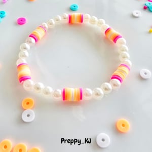 May include: A white pearl bracelet with colorful striped beads. The bracelet is on a white surface with small colorful circles scattered around it. The text "Preppy_KJ" is written on the surface.