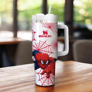 May include: A white and silver Stanley tumbler featuring Spider-Man graphics. The tumbler has a red spiderweb design and a large handle. The Spider-Man character is depicted in his classic red and blue suit. The tumbler includes a white straw.
