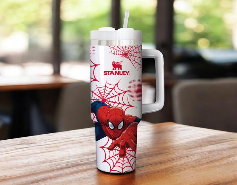 May include: A white and silver Stanley tumbler featuring Spider-Man graphics. The tumbler has a red spiderweb design and a large handle. The Spider-Man character is depicted in his classic red and blue suit. The tumbler includes a white straw.